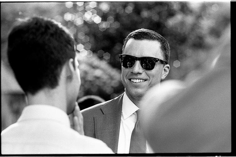 Nikon 85mm 1.5 bokeh out of focus points of light polygons sunglasses raybans clubmaster smile teeth man wedding cocktail hour reception social johnny martyr film b&w black and white portrait handsome